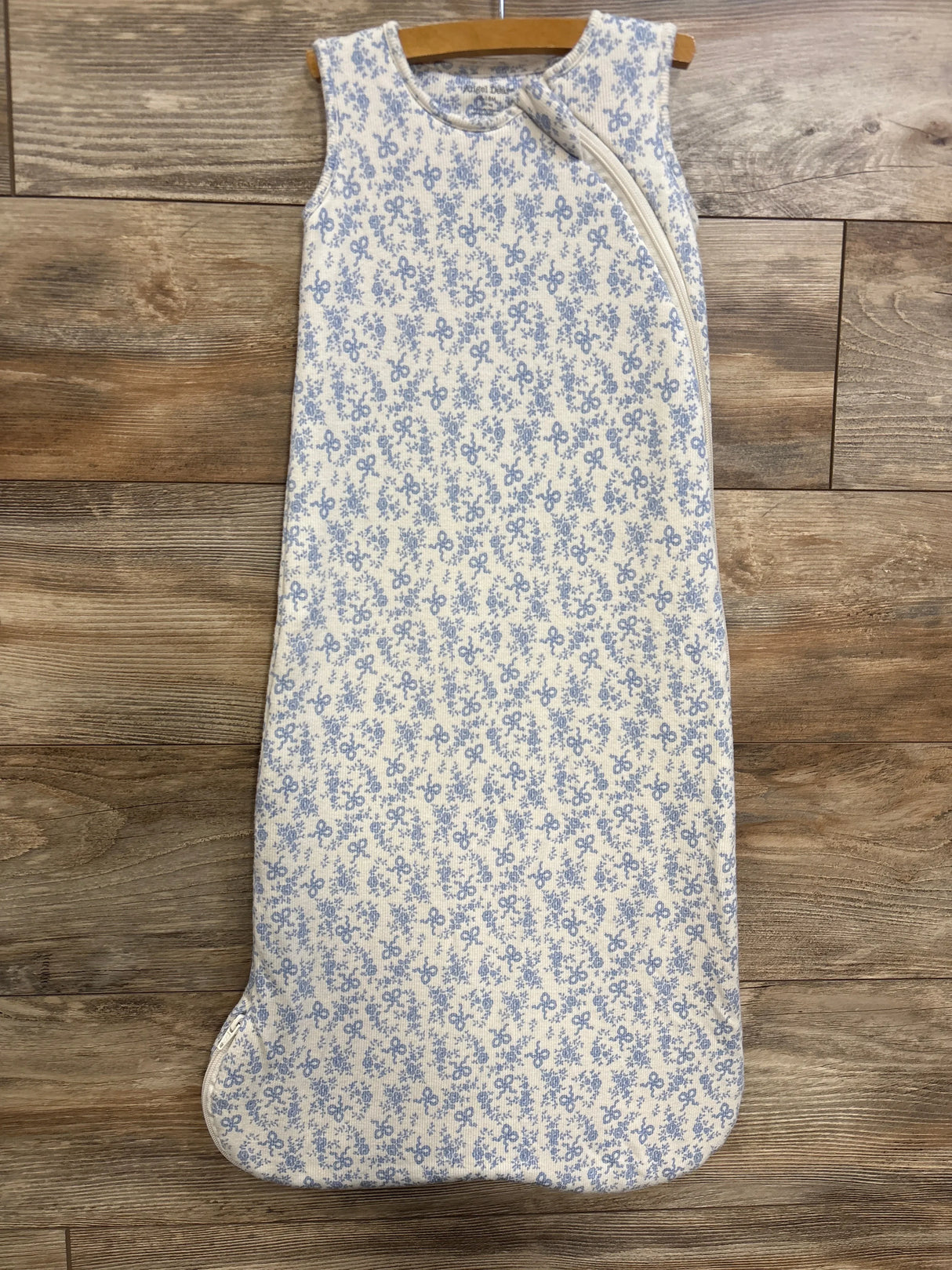 Front view of Angel Dear 1.0 TOG white sleep bag showcasing small blue floral-and-bow pattern and diagonal zipper, laid flat on wood floor