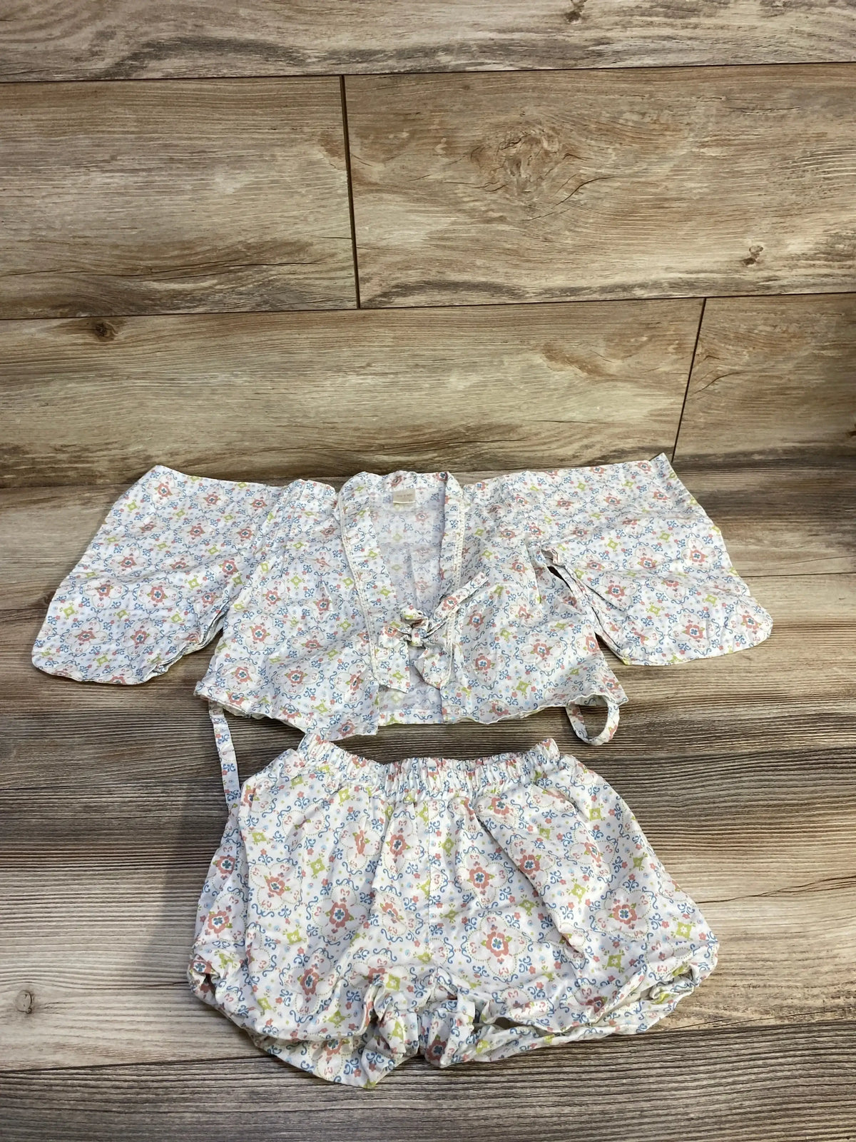 Flat lay of Tete a Tete floral wrap-style top and shorts set in white, showcasing floral print and cotton fabric on wood flooring