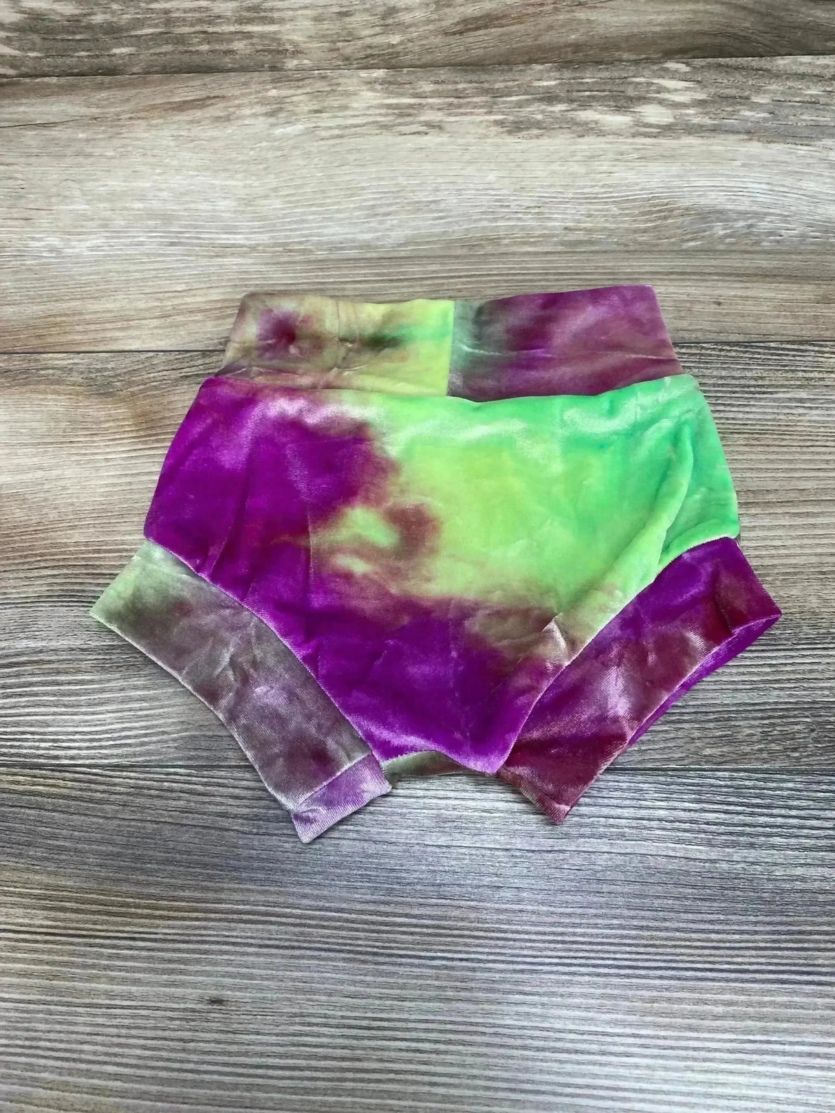 Flat lay of velvet baby bloomers in green and purple showing high waistband and cuffed legs on wood background