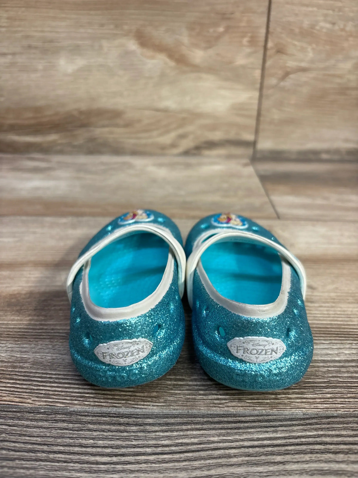 Front view of Crocs Frozen Mary Jane flats in blue glitter showcasing Frozen badges and white straps on a wood plank floor