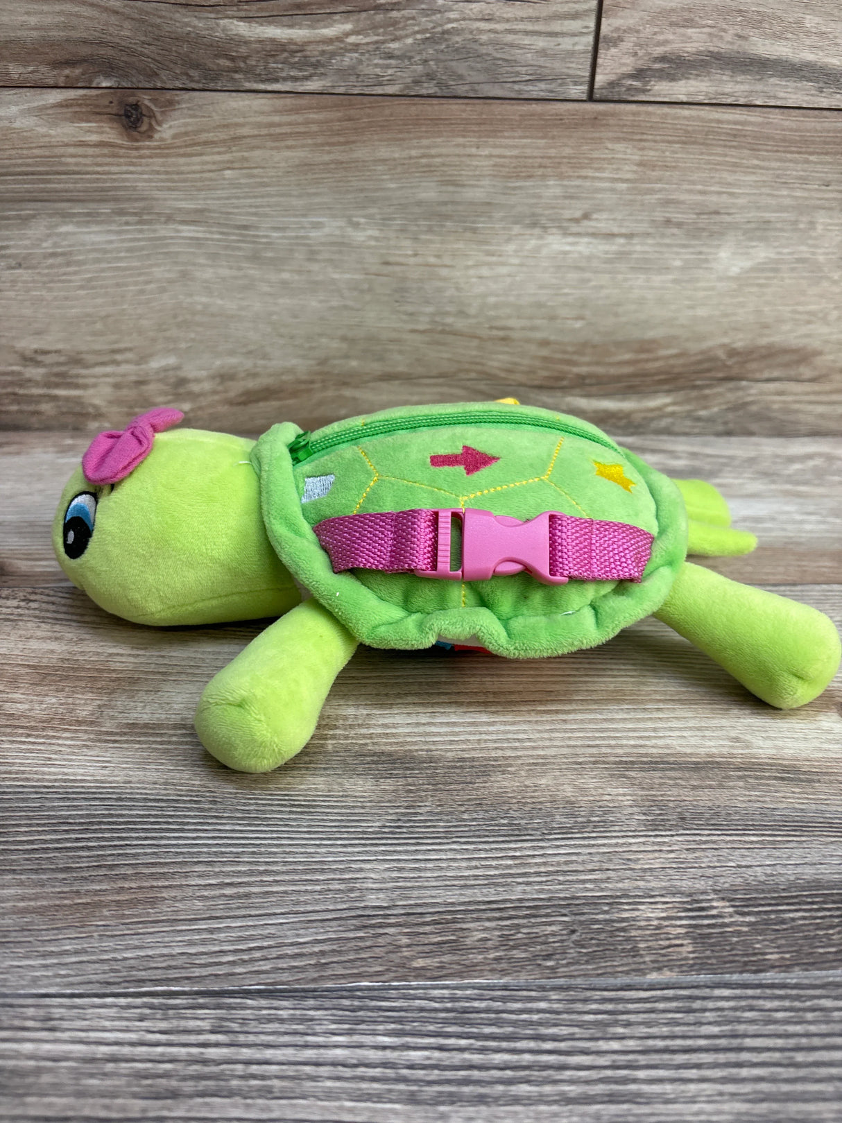 Plush green turtle toy with pink strap, zipper, and wooden background
