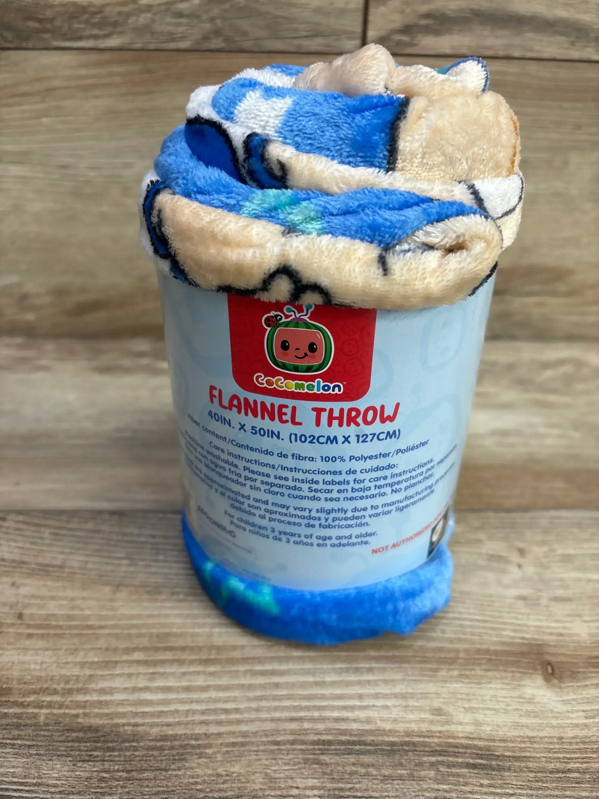 Front view of Cocomelon flannel throw rolled with blue plush fabric and label Flannel Throw 40in x 50in on wooden background