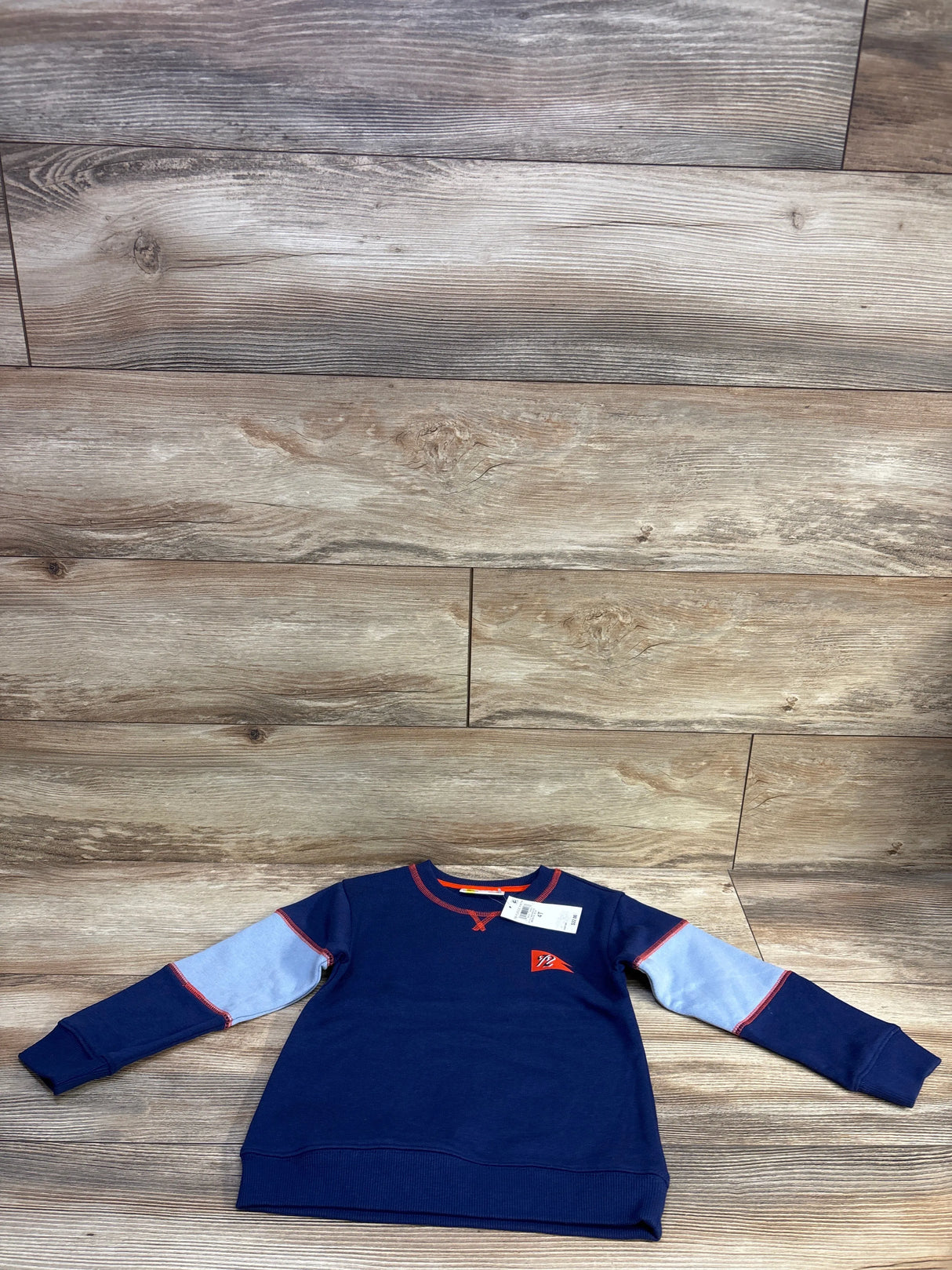 Front flat lay of navy toddler pullover sweatshirt with light-blue sleeve panels, red trim and chest pennant logo on wood background