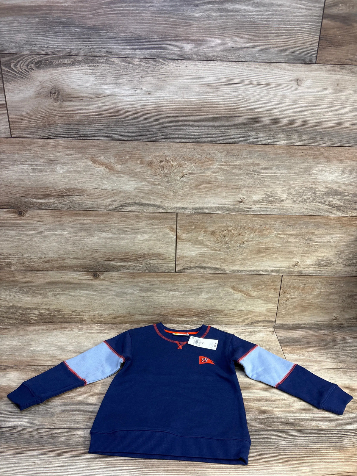 Front flat lay of navy toddler pullover sweatshirt with light-blue sleeve panels, red trim and chest pennant logo on wood background