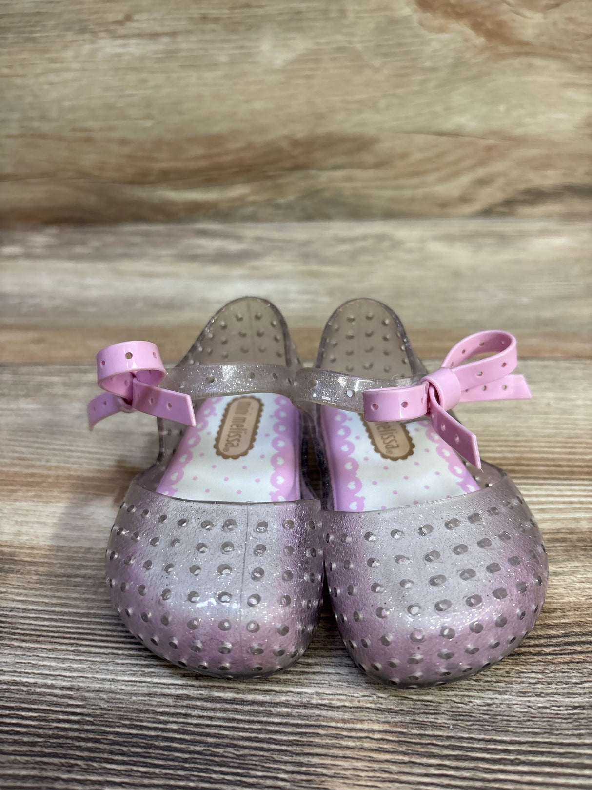 Front view of Mini Melissa Ultragirl jelly Mary Jane shoes showcasing silver glitter finish and pink bows on a wooden background