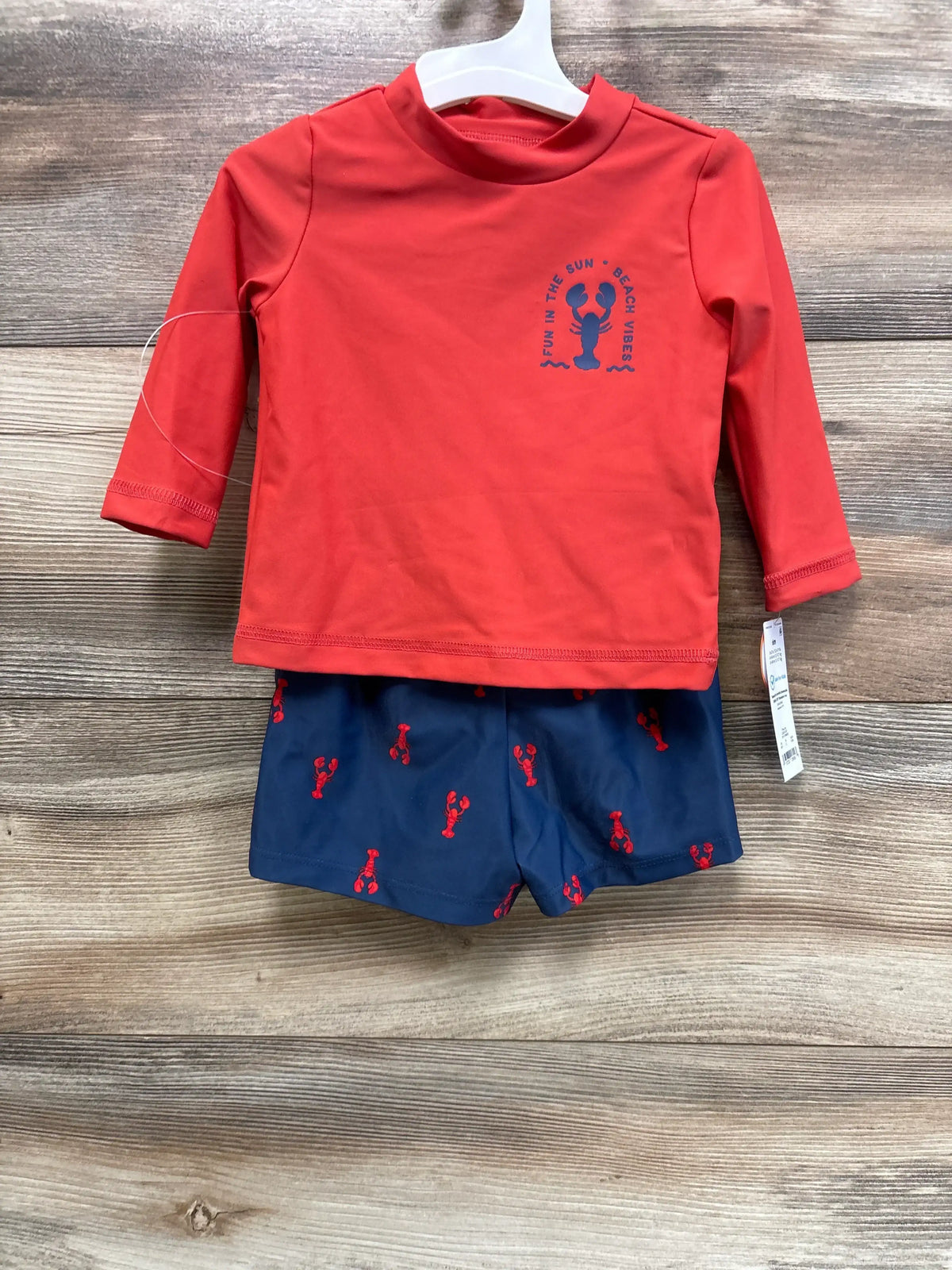 Front of baby 2-piece swim set, red rash top with lobster graphic and navy trunks with lobster print, on white hanger on wood