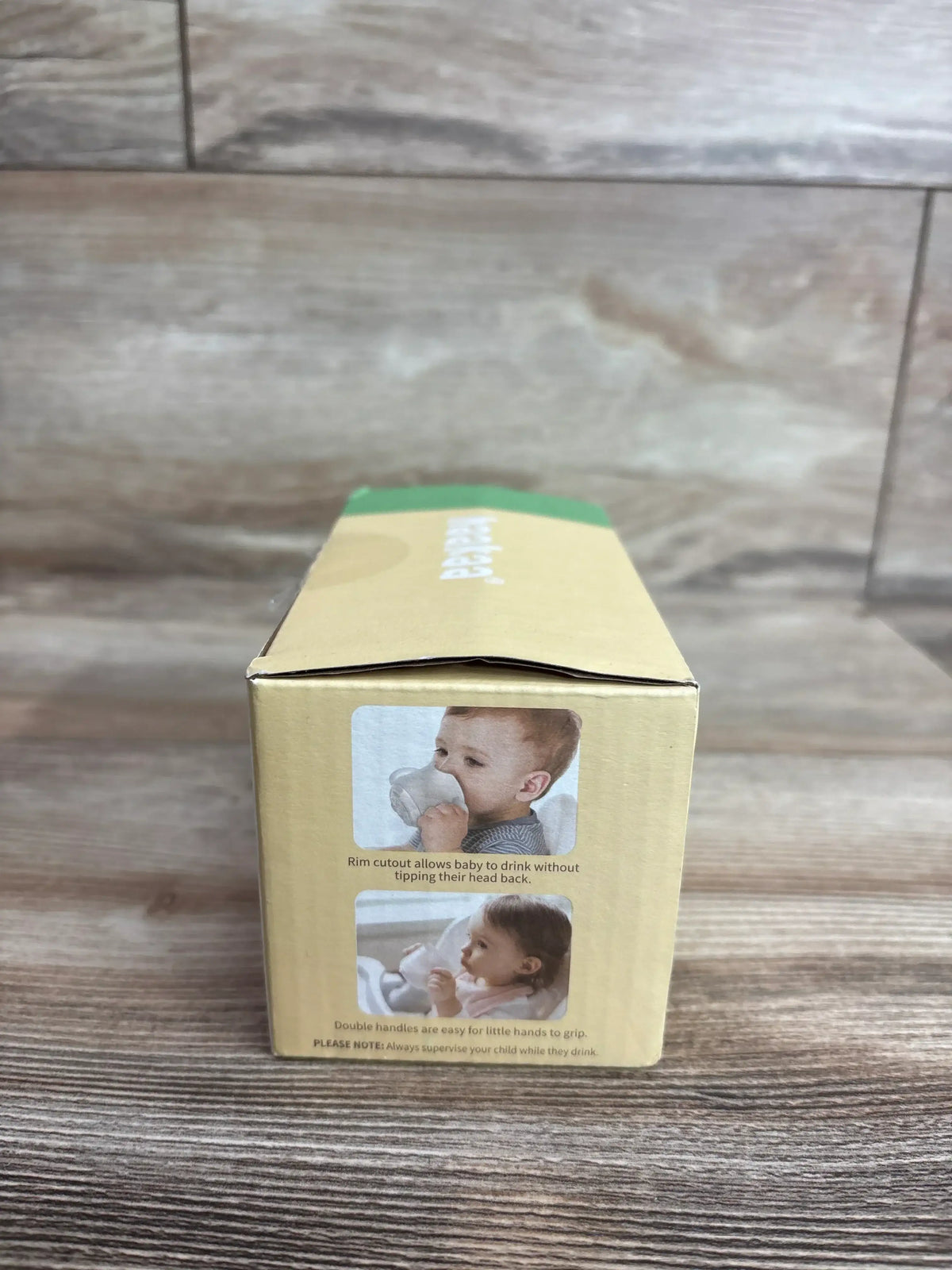Front view of Haakaa silicone drinking cup box showing baby photos and text about rim cutout and handles on wood surface