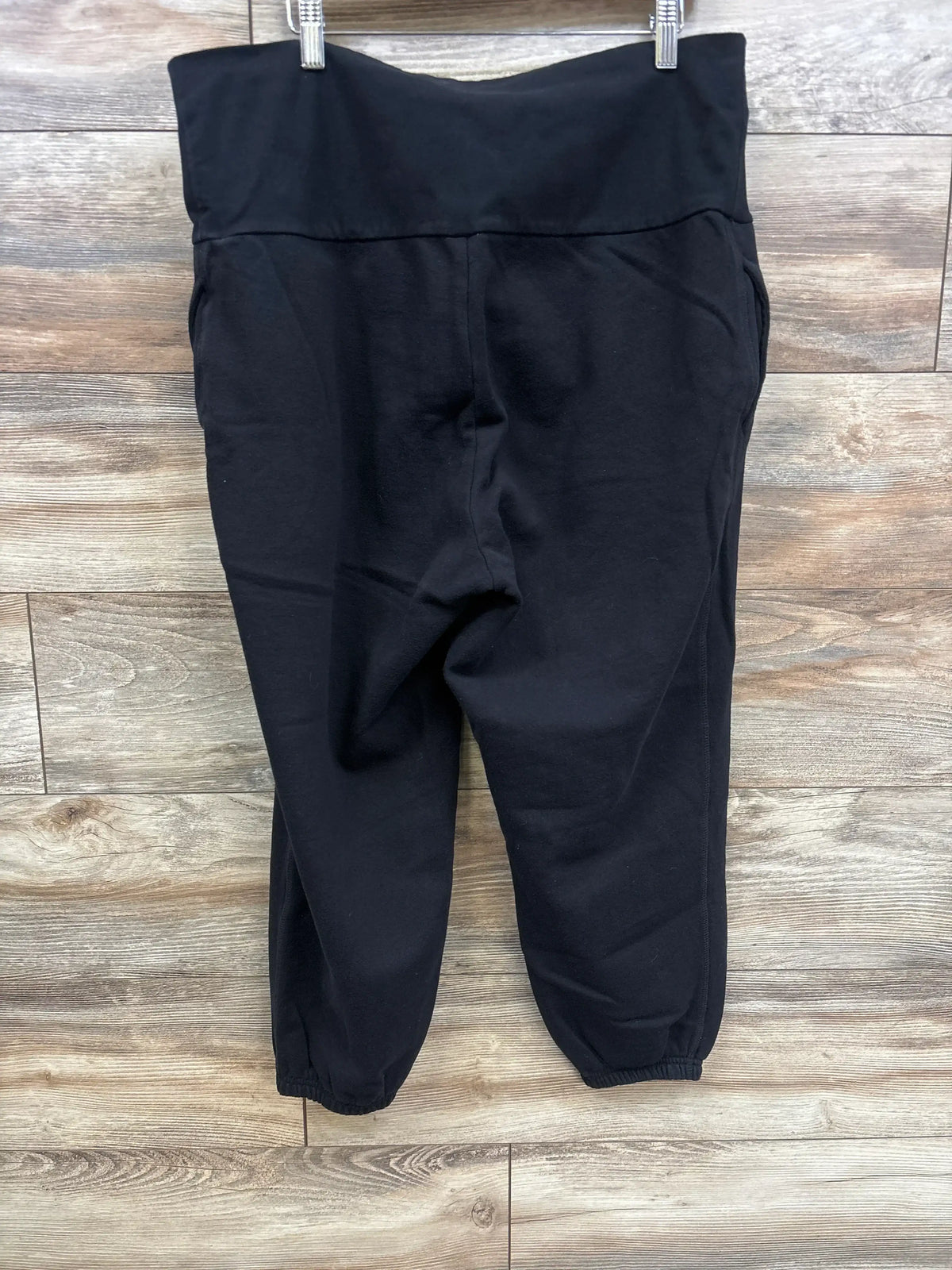 Front view of black maternity joggers with rollover waist, side pockets, tapered legs and elastic cuffs on wood background
