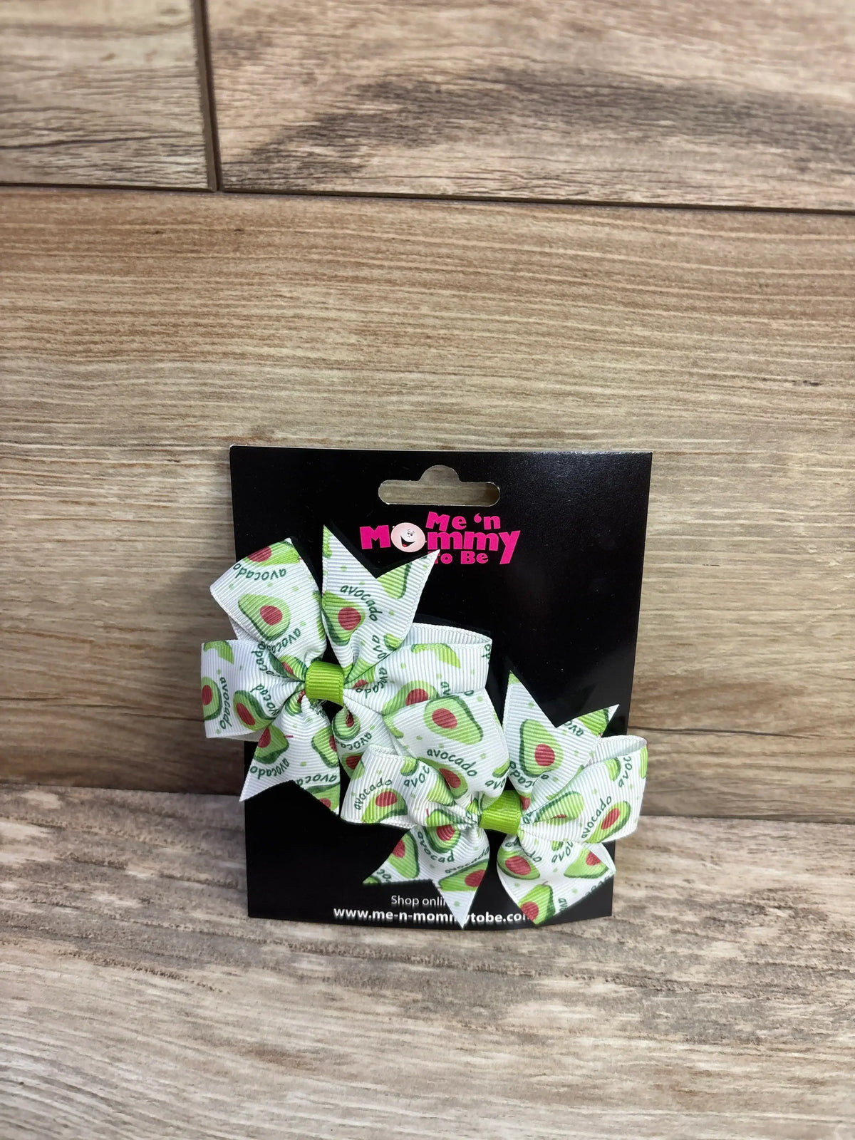 Front view of two fanned-out avocado-print hair bows 2-pack on black Me n Mommy To Be display card against wood background