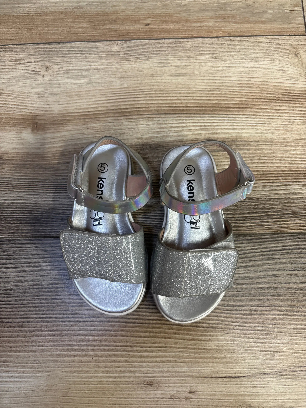 Top view of Kensie Girl glitter sandals in silver showcasing glitter finish and ankle straps with a wood background