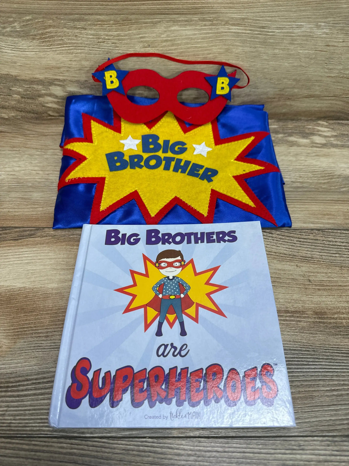 Front view of a Big Brother superhero costume set with red mask, blue costume, and matching book on a wood floor
