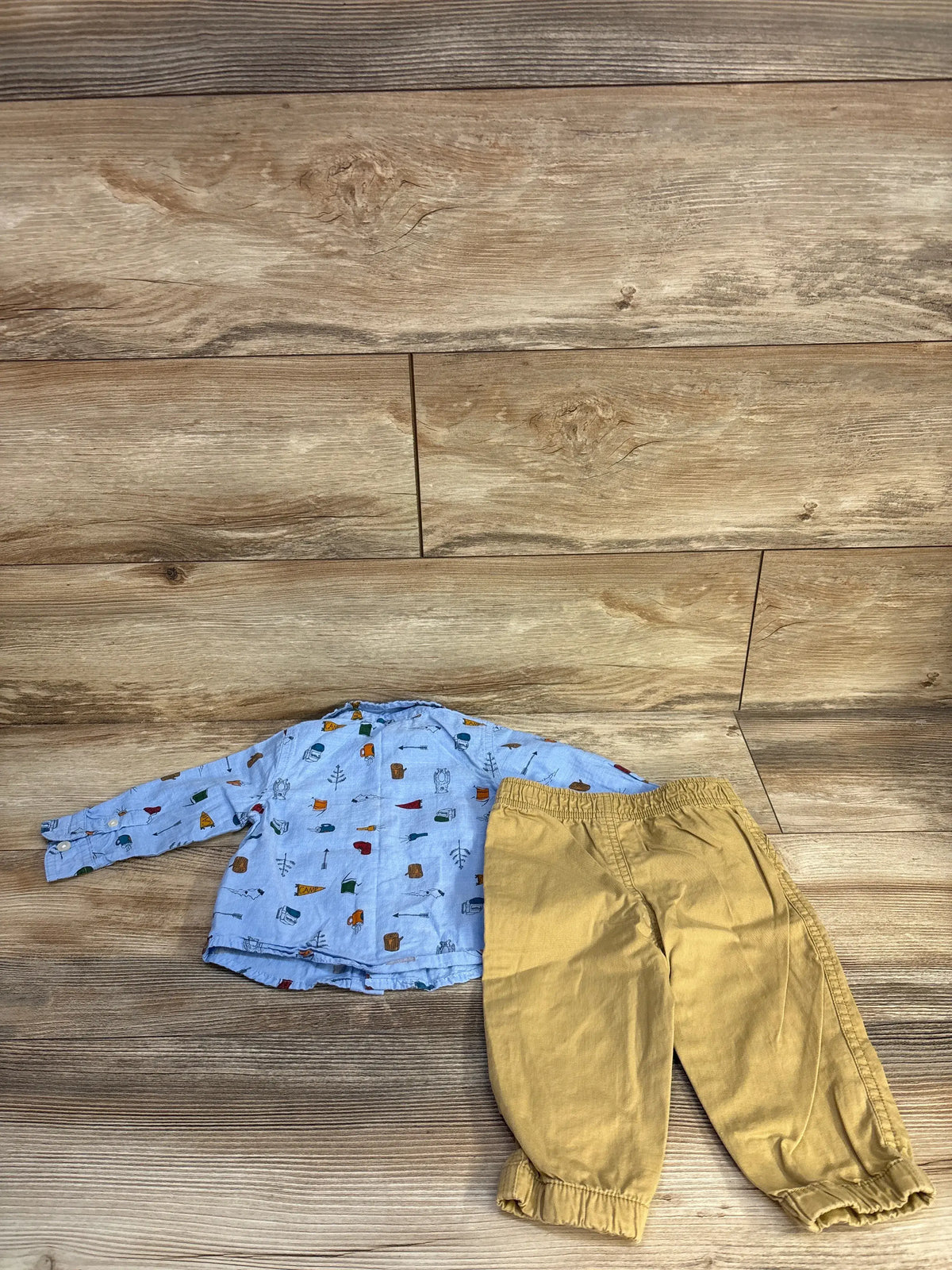 Flat lay of Baby B.U.M two-piece camping set showing a blue patterned button-up shirt and tan pants on wood