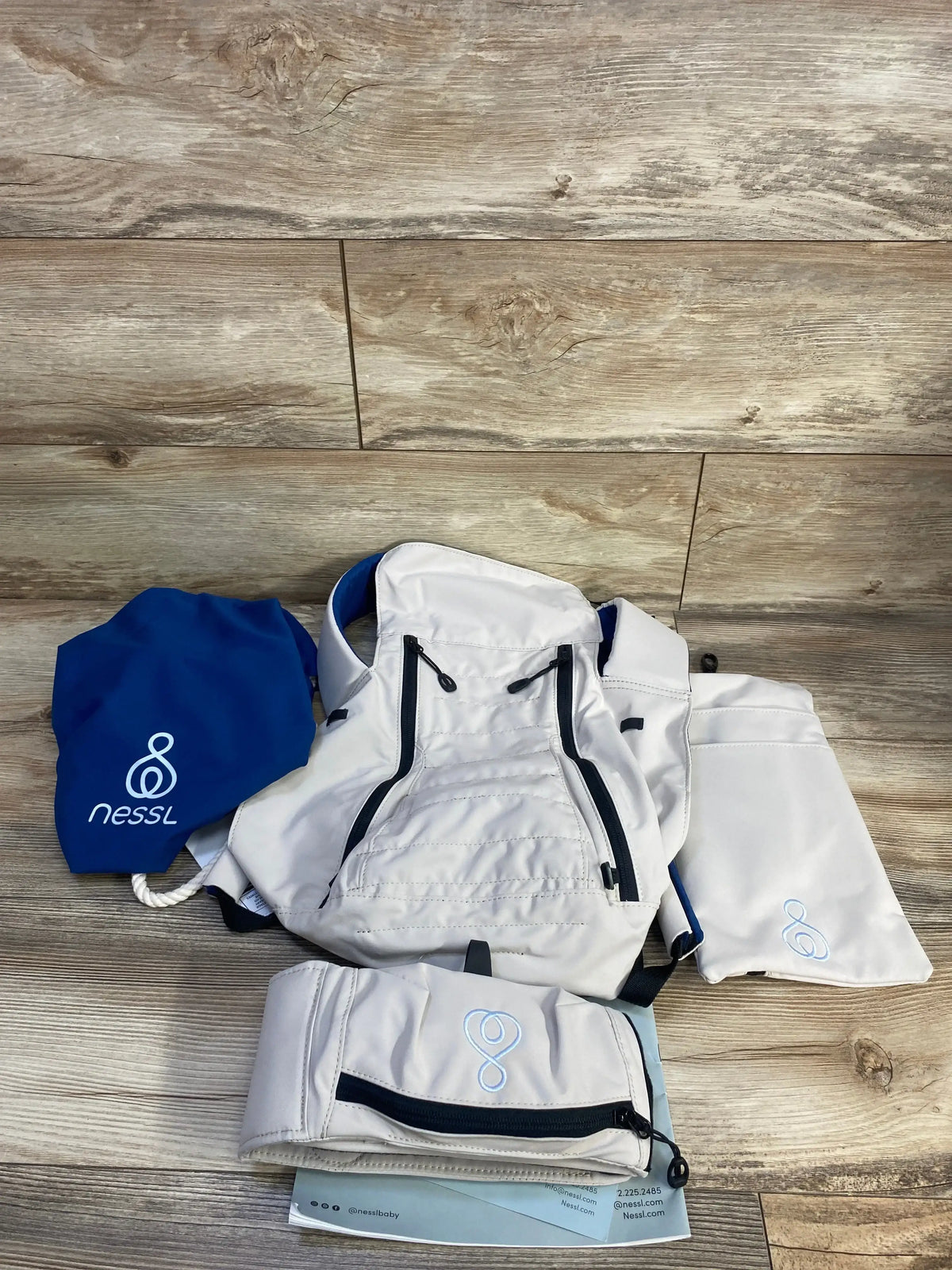 Flat lay of NWT Nessl baby carrier in oatmeal with navy blue drawstring storage pouch and instruction booklet on wood plank floor