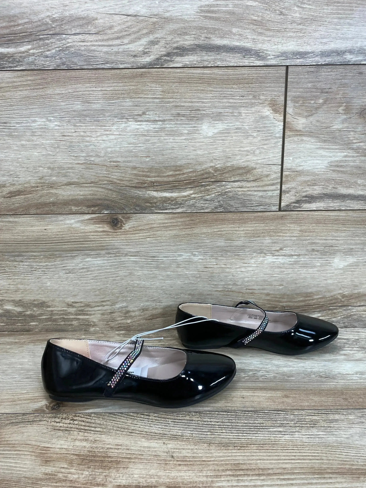 Side view of girls' black faux patent leather ballet flats showcasing jeweled Mary Jane strap on light wood floor