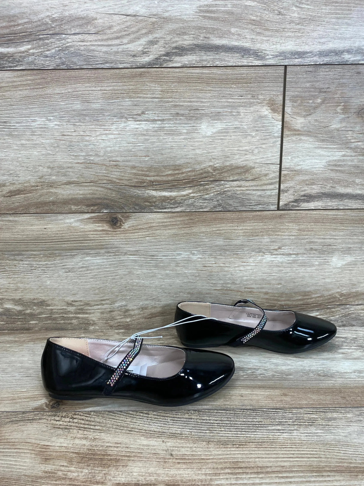Side view of girls' black faux patent leather ballet flats showcasing jeweled Mary Jane strap on light wood floor