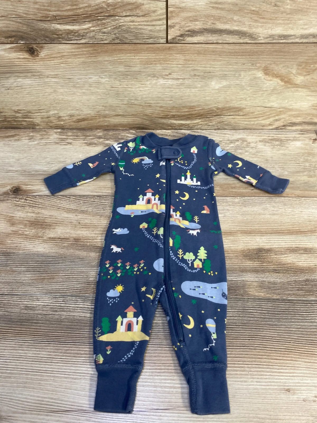 Front flat-lay of blue Hanna Andersson sleeper showcasing zipper front, fold-over cuffs, and castle-and-moon print on wooden background