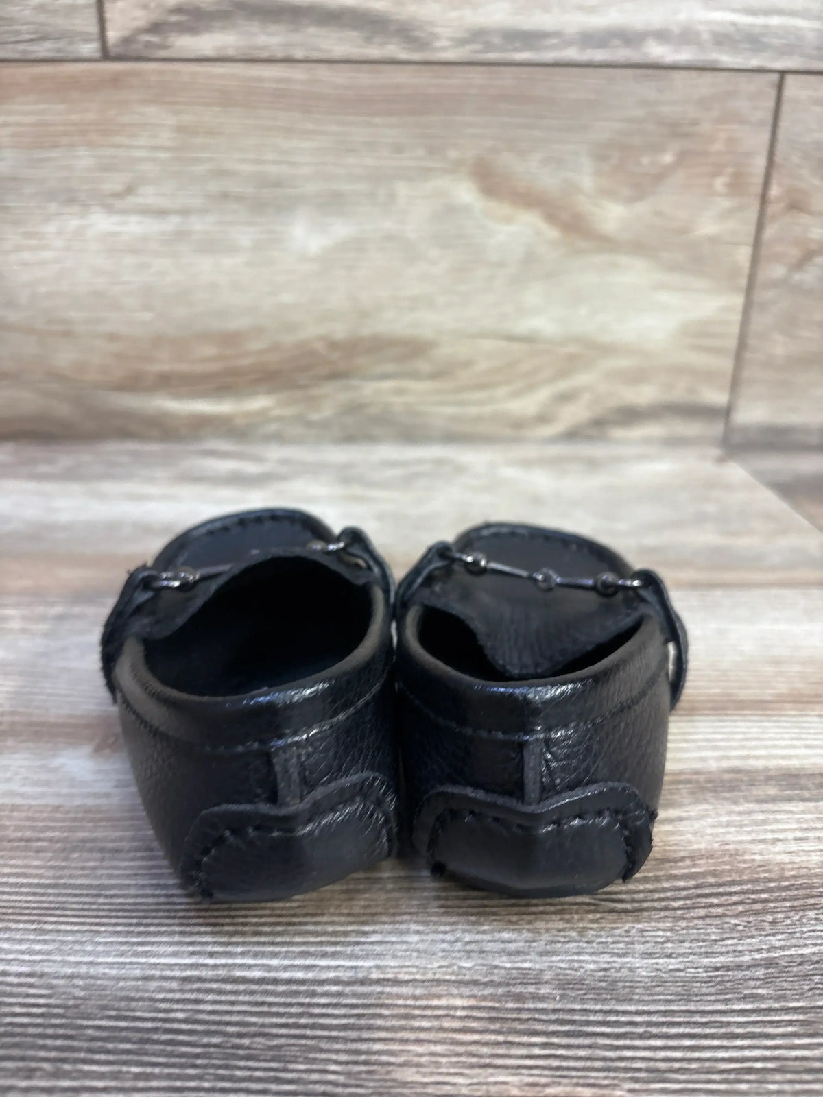 Rear view of Minibella black soft leather slip-on baby boat shoes showcasing stitched heel and pebble leather texture on woodgrain surface