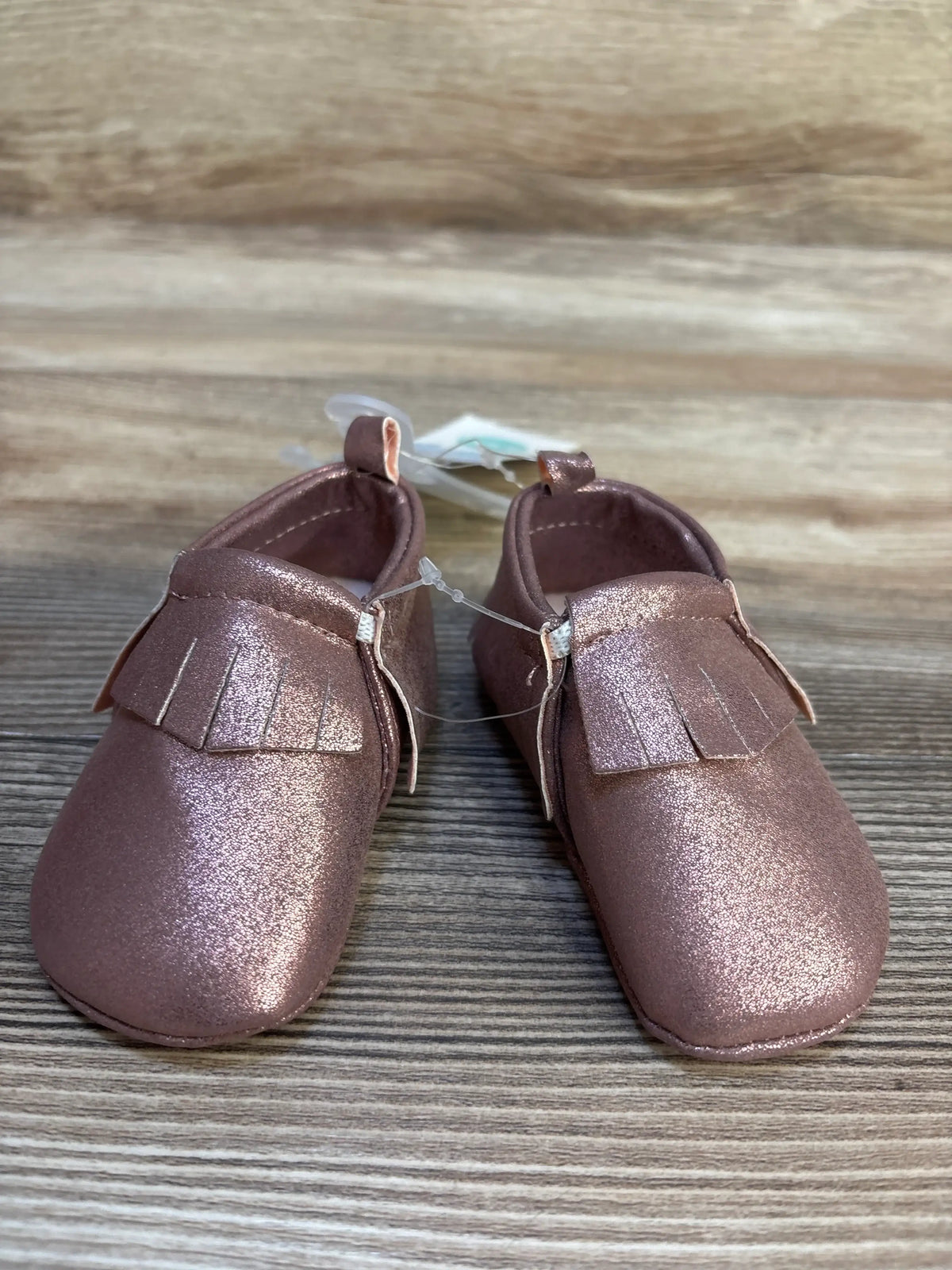 Metallic pink baby moccasins with fringe detail on wooden surface