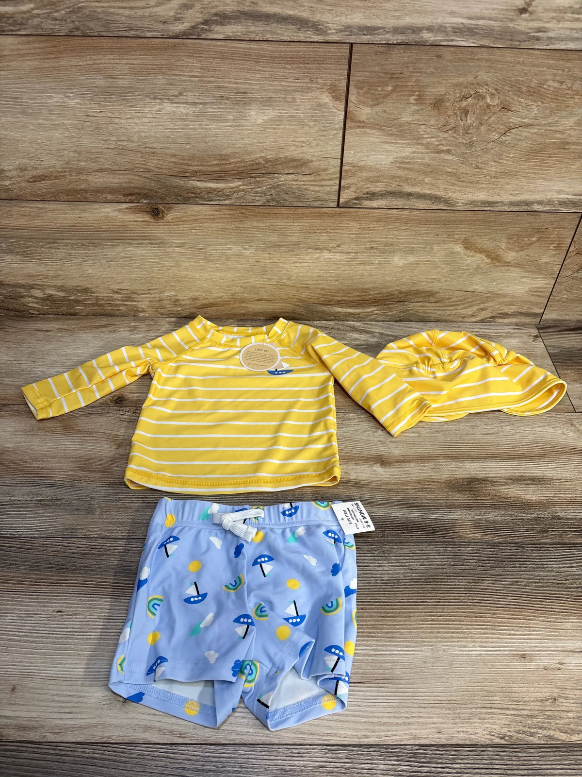 Top view of First Impression three-piece swim set showcasing yellow striped rash guard and hat with blue printed trunks