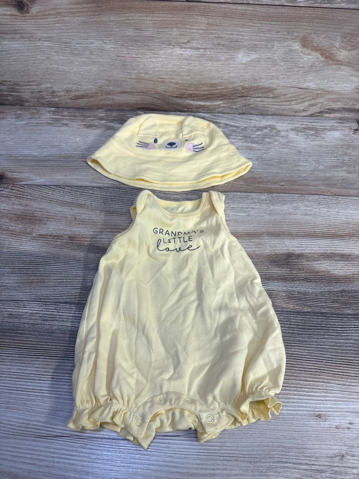 Front view of yellow romper and hat set showcasing ruffled trim, cat-face hat, and Grandma’s Little Love lettering on wood
