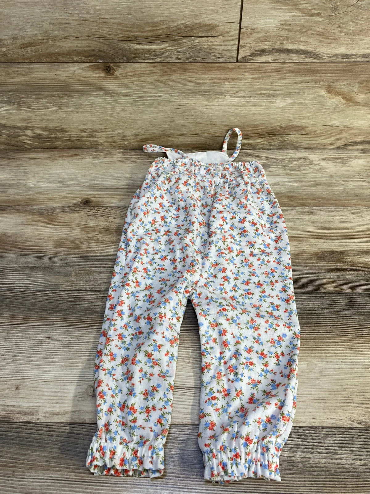 Front flat lay of white floral toddler sleeveless jumpsuit showing thin shoulder straps and elastic ruffle ankle cuffs on wood background