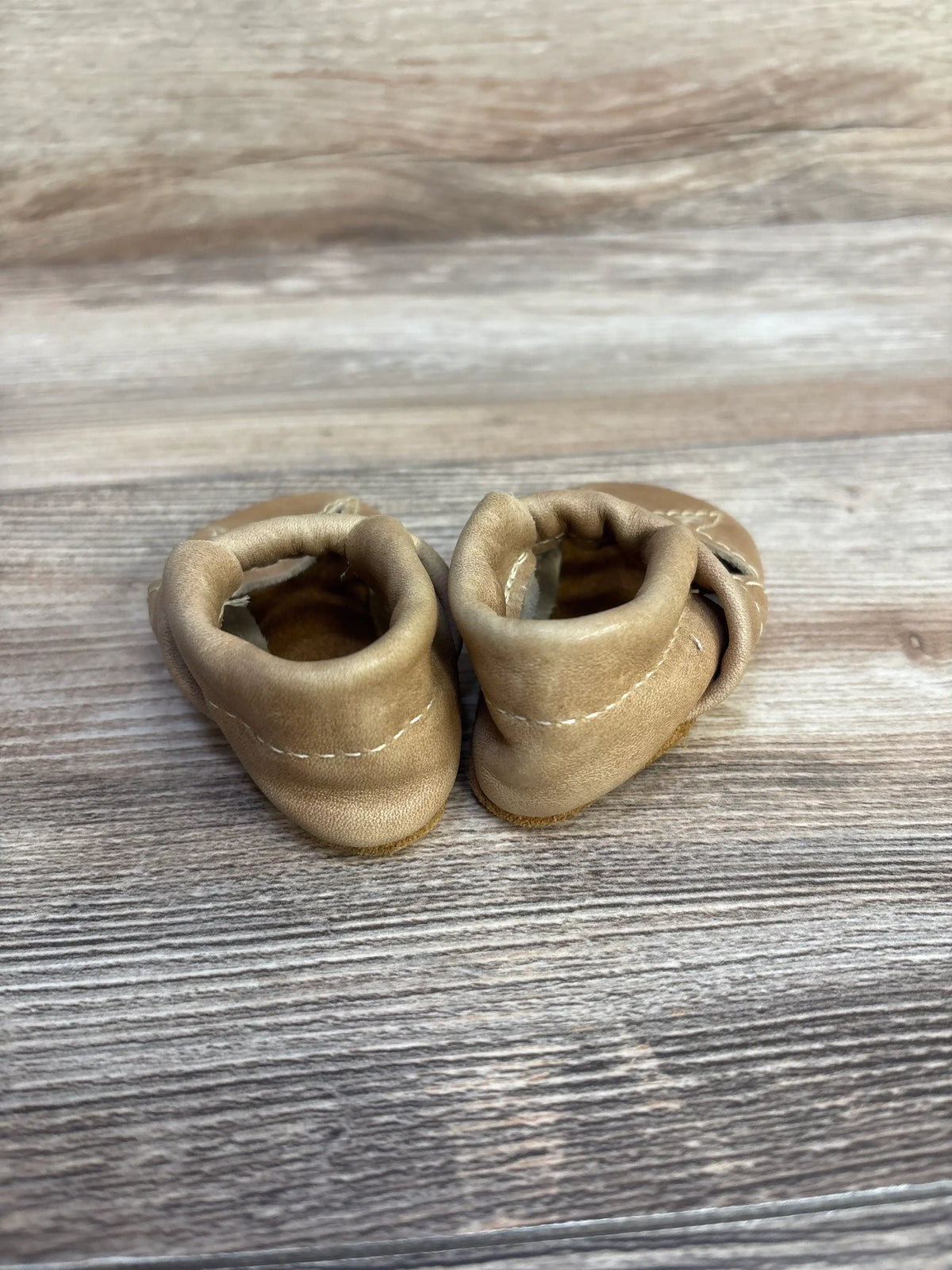 Rear view of newborn tan leather baby moccasins showing elasticized ankle, stitching, and suede soles on light wood background