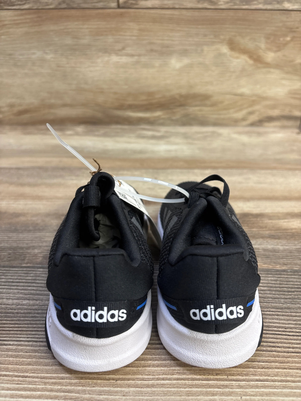 Rear view of kids’ Adidas Racer TR 2.01 sneakers in black, size 9C, showing Adidas logo on heels and white soles on wooden background