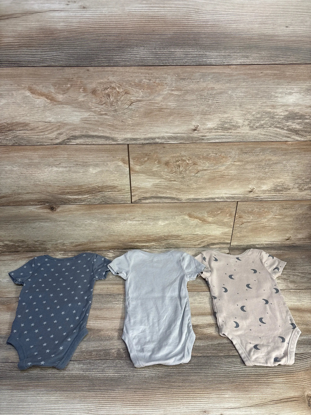 Front view of Cloud Island 3-pack bodysuit set showcasing solid grey, star, and moon prints on a wood floor