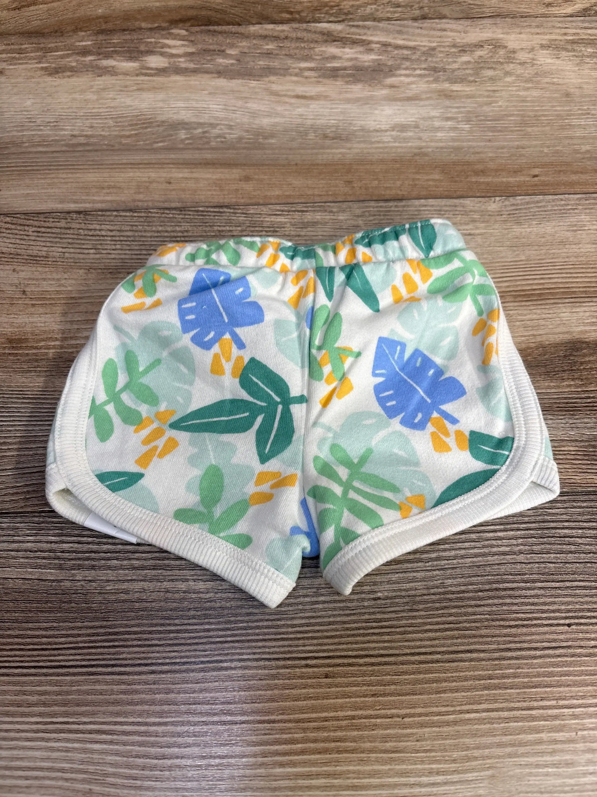 Top-down view of Jumping Beans white baby shorts showing green, blue and yellow leaf print, elastic waistband and white ribbed trim on wood surface