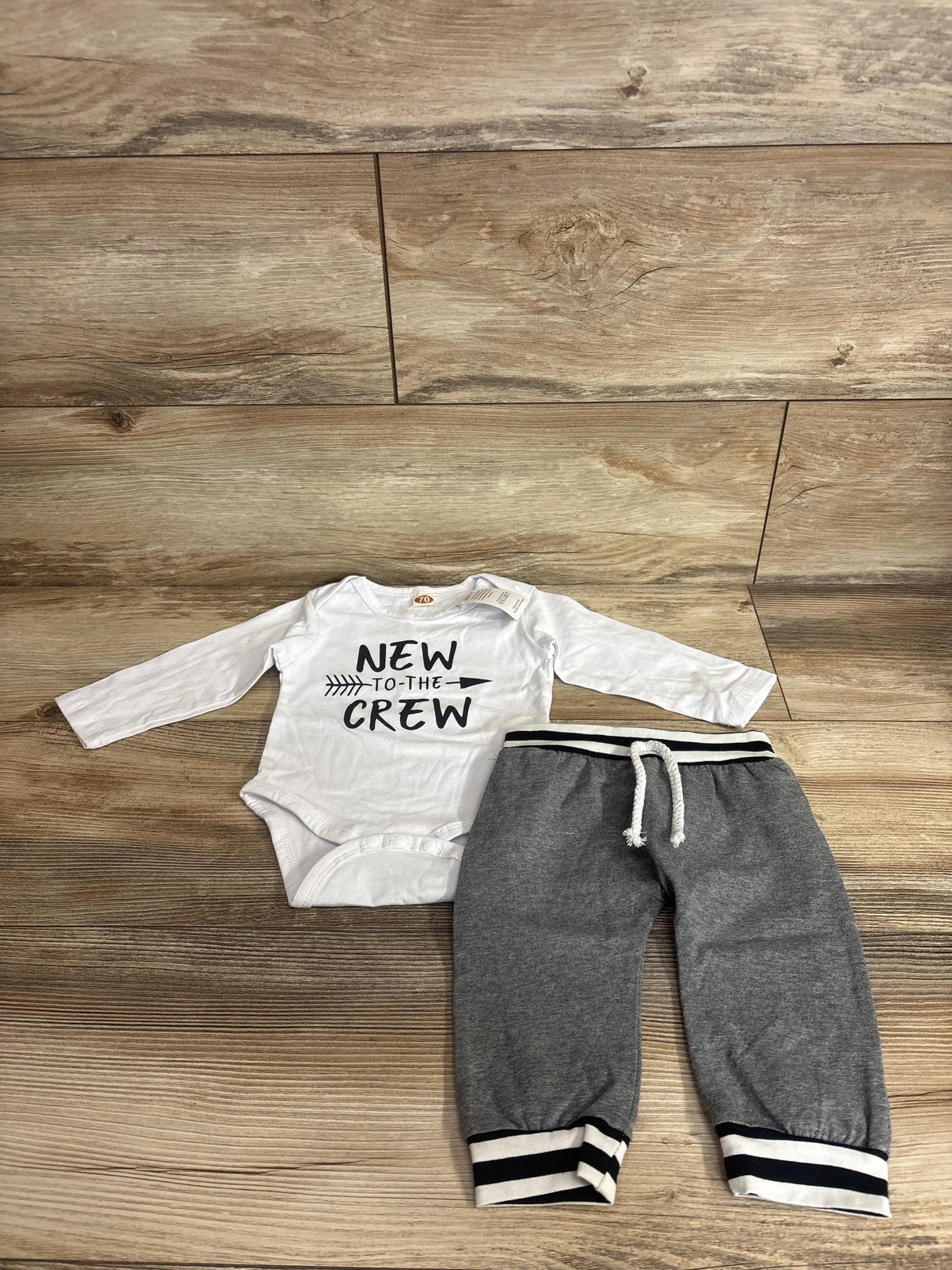 Flat lay of white bodysuit and gray joggers showing NEW TO THE CREW text and drawstring striped cuffs on wood floor