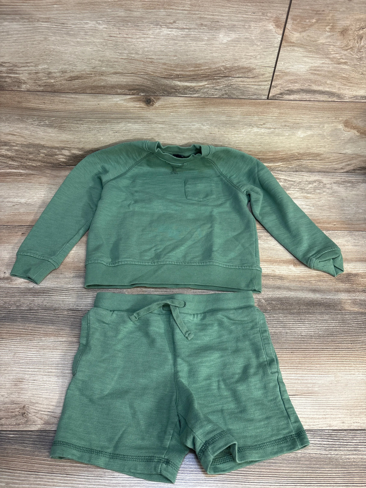 Flat lay of Janie & Jack green sweatshirt and shorts size 5T showing chest pocket and drawstring waist on wood background