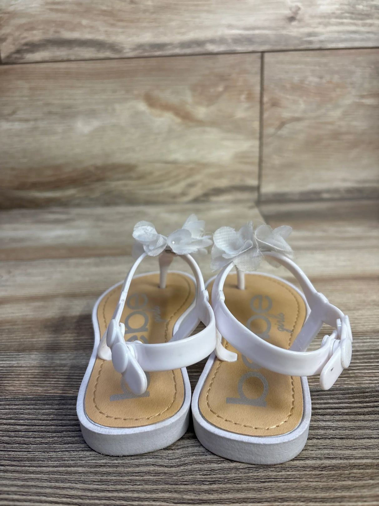 Front view of white Bebe toddler chiffon flower sandals showing flower accents and buckled ankle straps on wood background