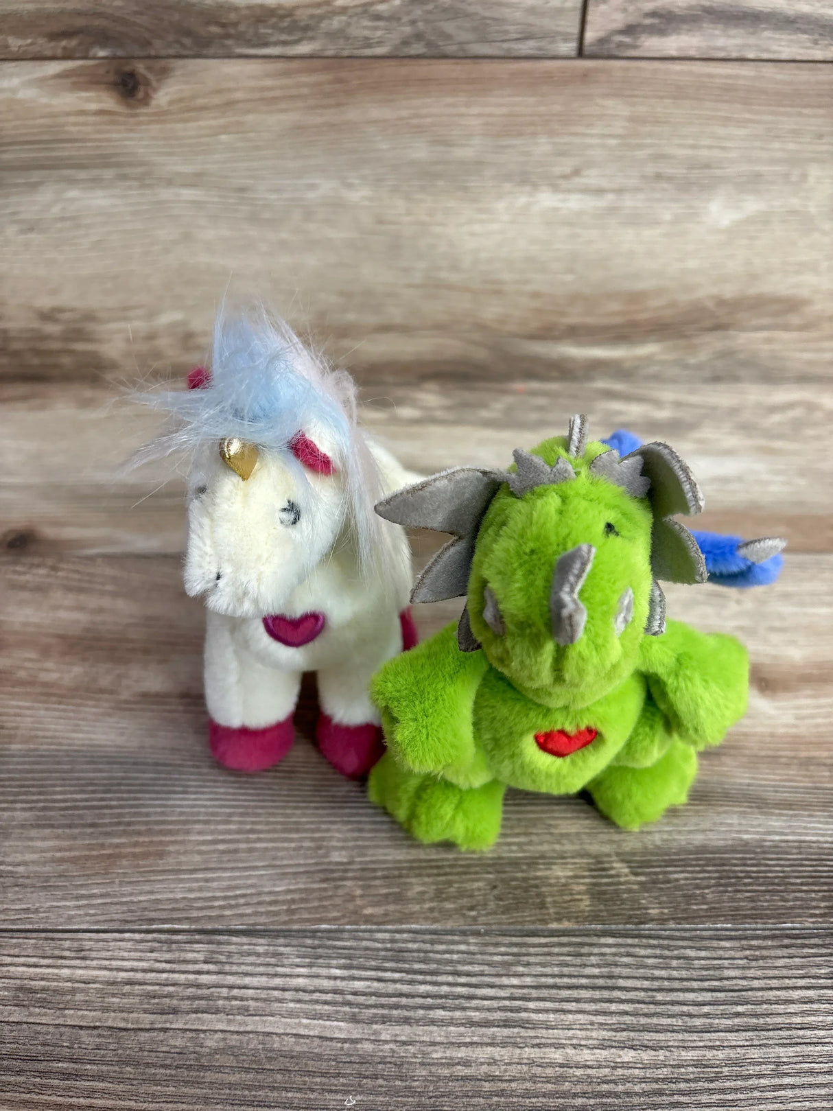 Two small plush toys on wood surface, one white unicorn with pink hooves and blue mane, one green dragon with wings