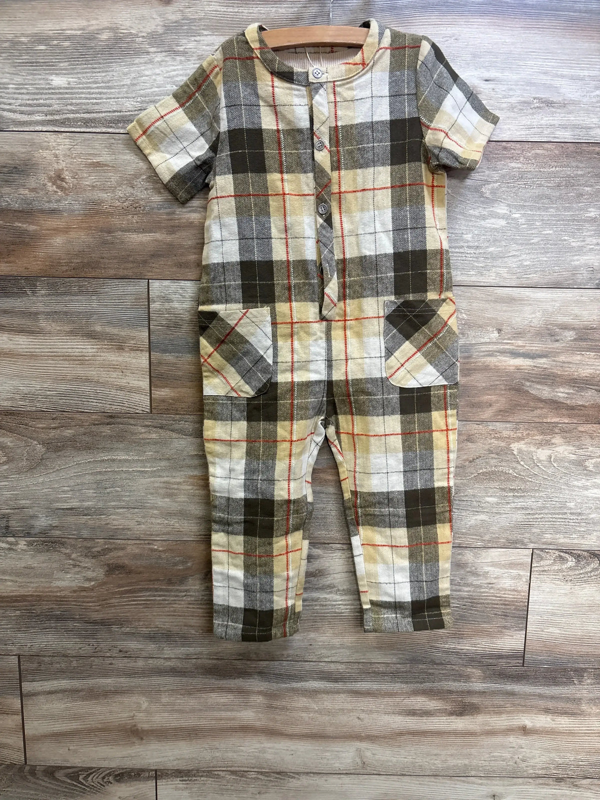 Flat-lay of beige and olive plaid kids jumpsuit showcasing short sleeves, front button placket and patch pockets on wood floor