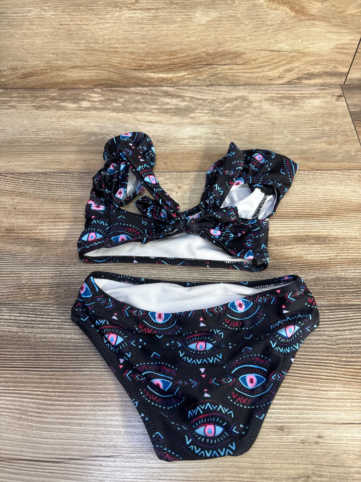 Top-down view of black two-piece toddler swimsuit with pink and blue evil eye print, knotted front top and matching bottoms on wood background
