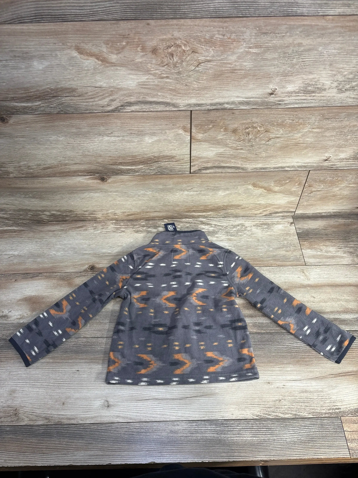 Child's long-sleeve fleece jacket with orange and white patterns on gray fabric laid flat on wooden floor