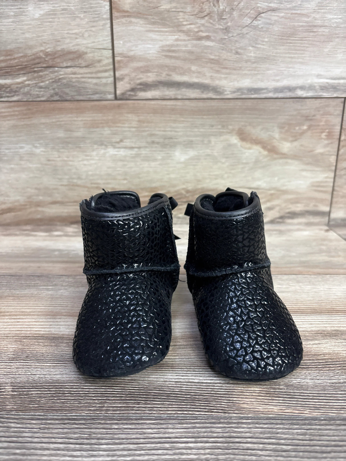 Front view of Ugg Jesse Bow II baby boots in black with textured pebble exterior, plush lining, and back bows on wood floor