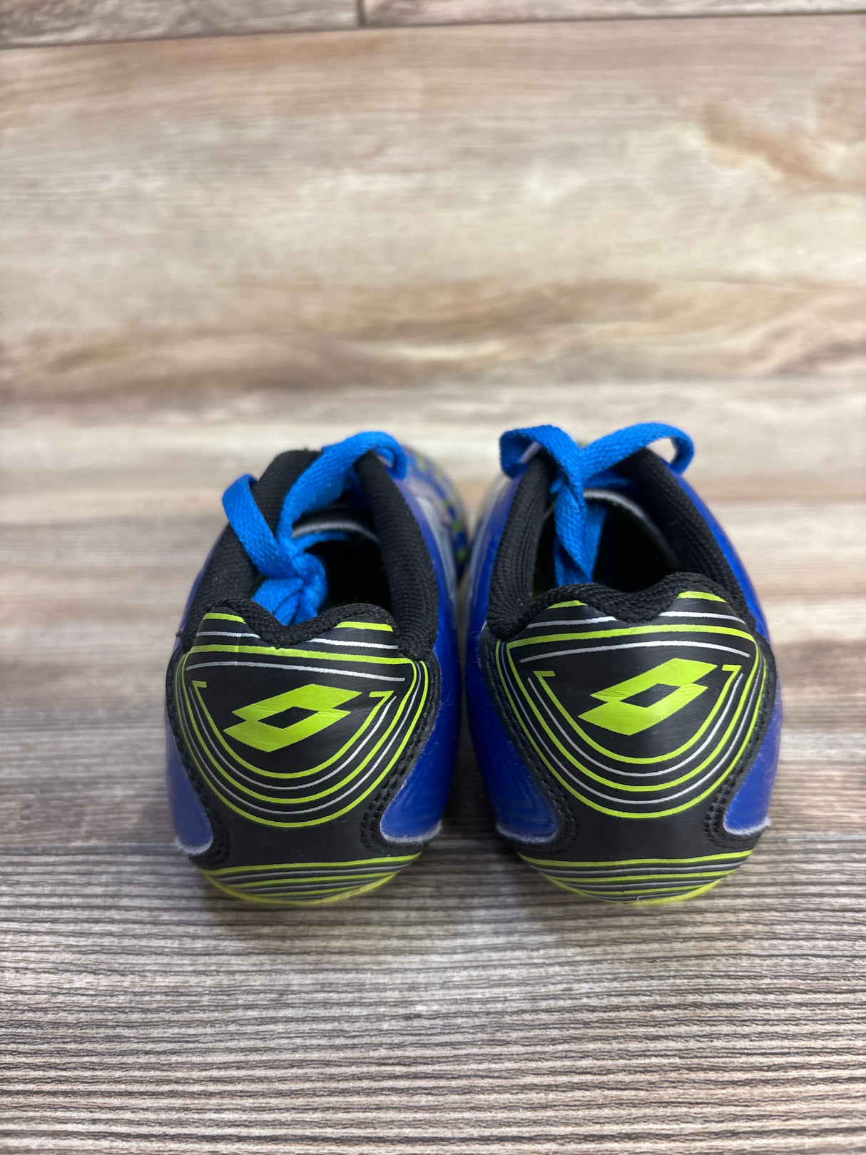 Rear view of Lotto Forza II Jr soccer cleats showcasing neon green Lotto logo accents and blue laces on a wood-grain tabletop
