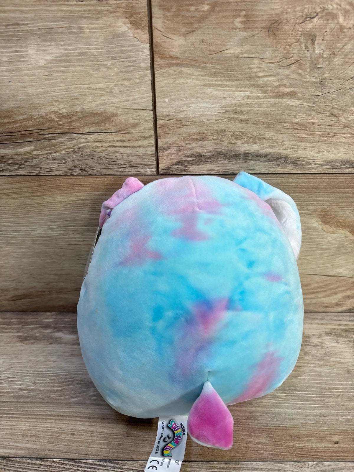 Back view of 8-inch Ryder the Bunny Squishmallows plush showing pastel blue-pink tie-dye fabric, small tail and ears on wooden plank background