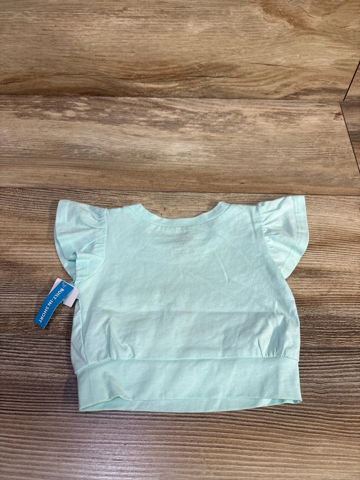 Back view of teal baby shirt showcasing flutter ruffle sleeves and elasticized hem on wood floor