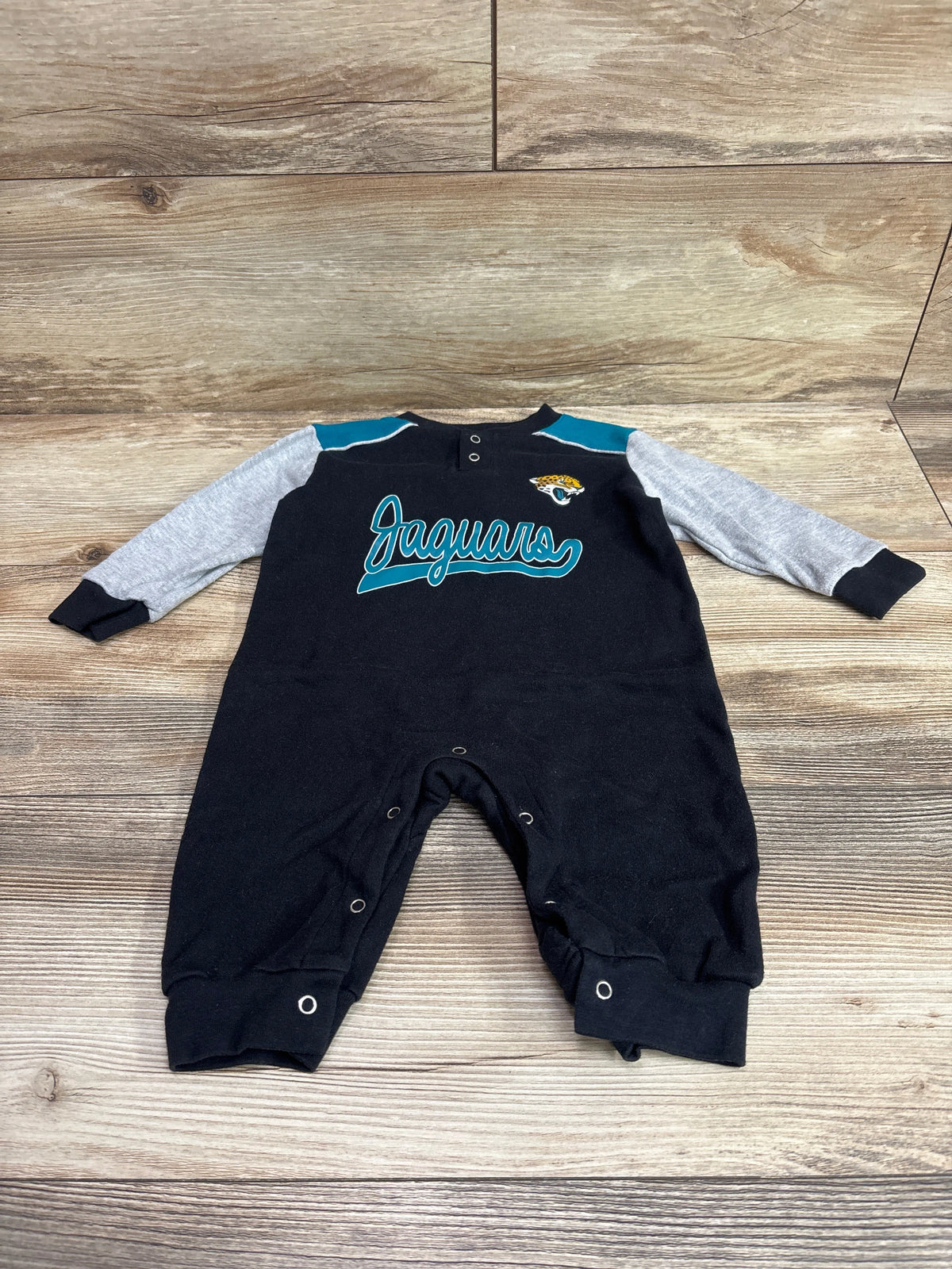 Front view of black Jaguars coverall showcasing gray sleeves, teal shoulder panels, and snap closures on a wood surface