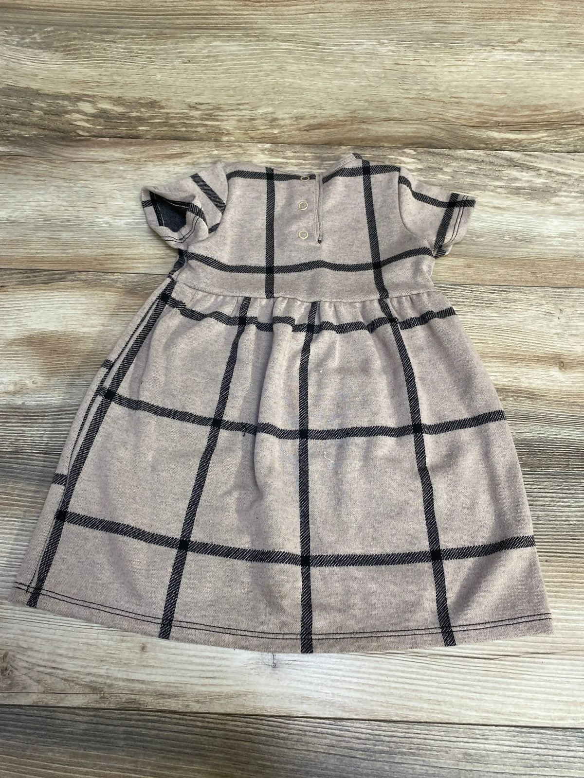 Flat lay of brown short-sleeve baby dress with black windowpane checks showcasing gathered waist and two-button placket on wood background