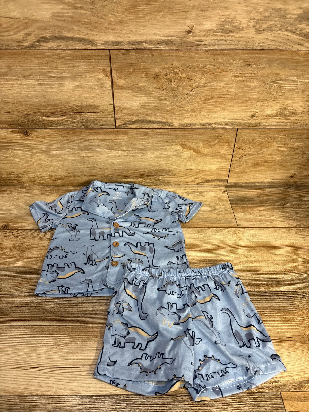 Top-down view of Carter’s blue two-piece coat-style pajamas with dinosaur print, button-front top, and bottoms on wood floor