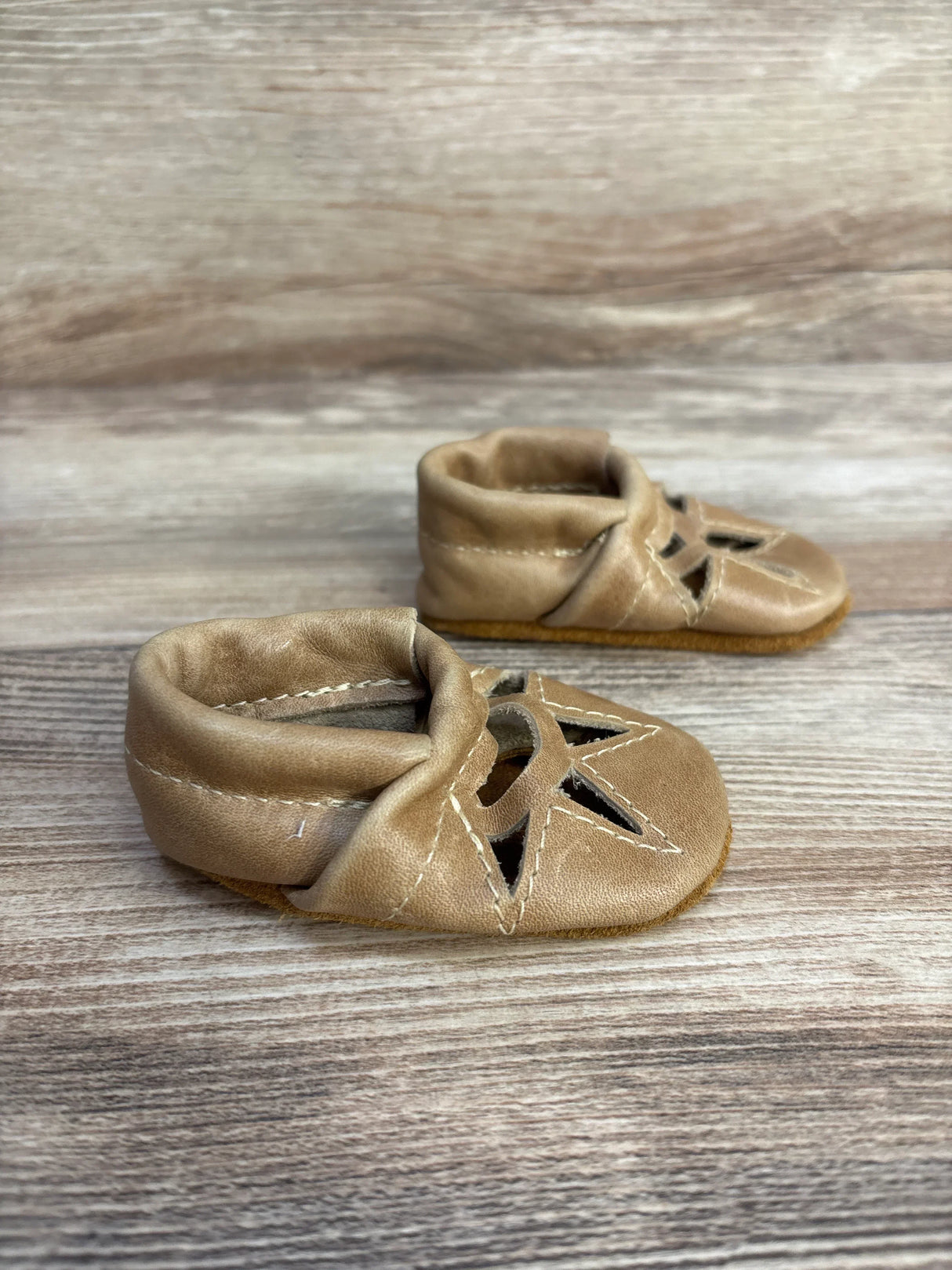 Three-quarter view of tan baby soft sole leather moccasins in newborn size showcasing star cutout details on wood plank floor