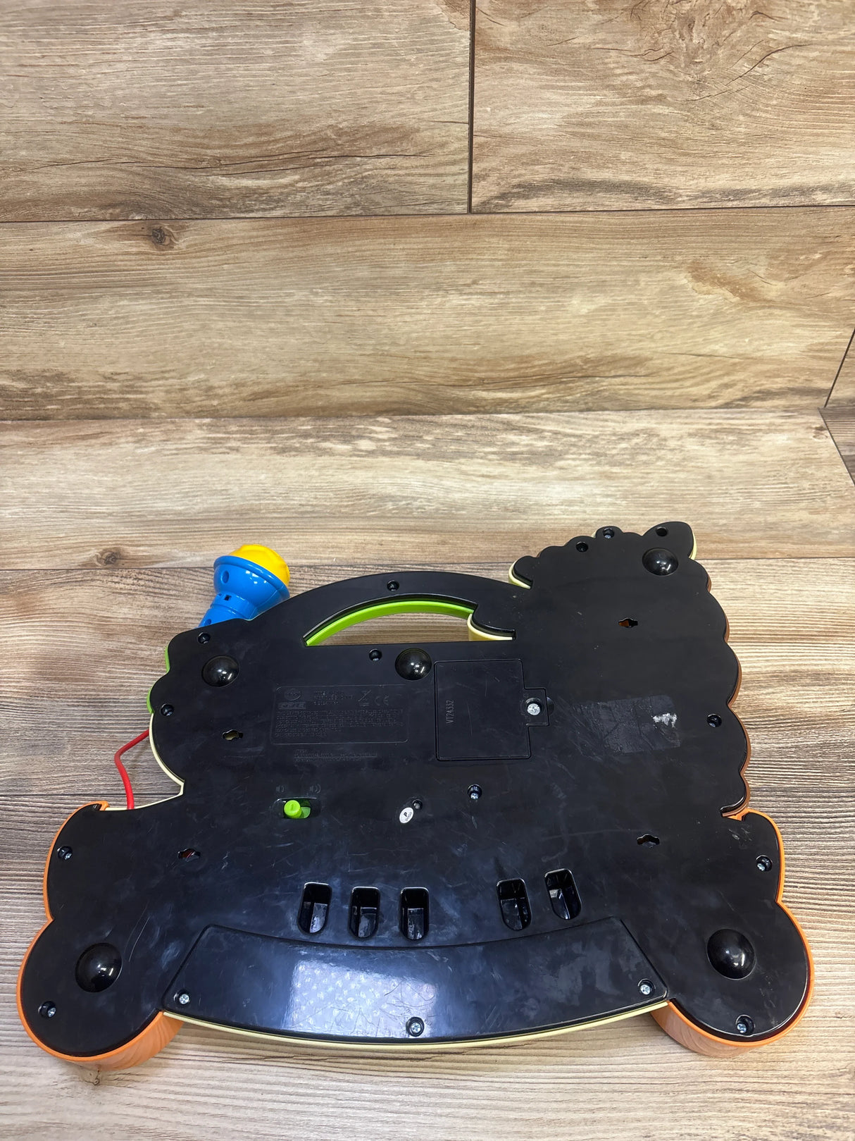 Bottom view of VTech Zoo Jamz giraffe piano showing battery compartment, green power switch, and attached blue microphone on wood background
