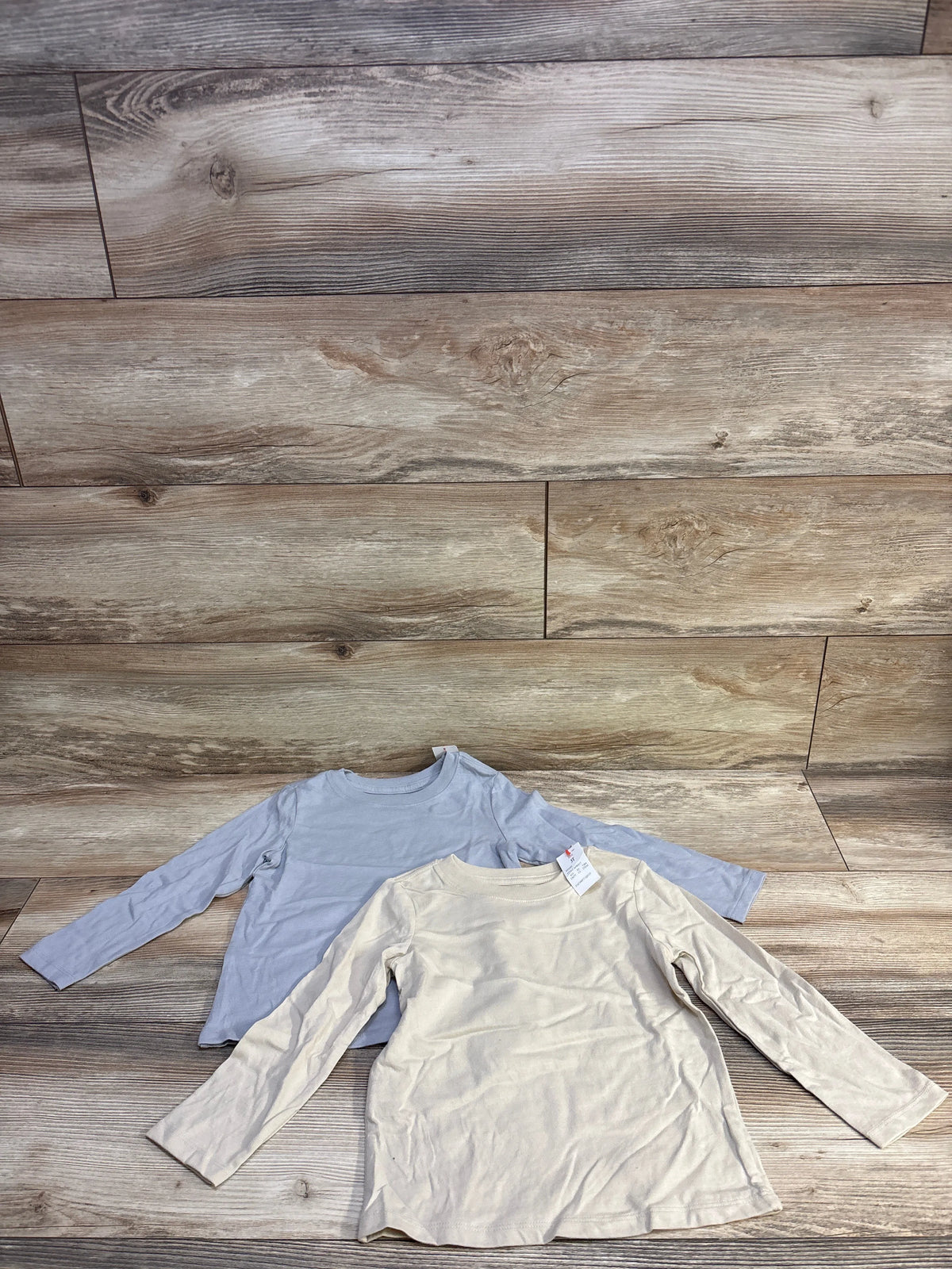 Flat lay of two toddler long-sleeve shirts (beige and light blue) showing attached tag on wood plank background