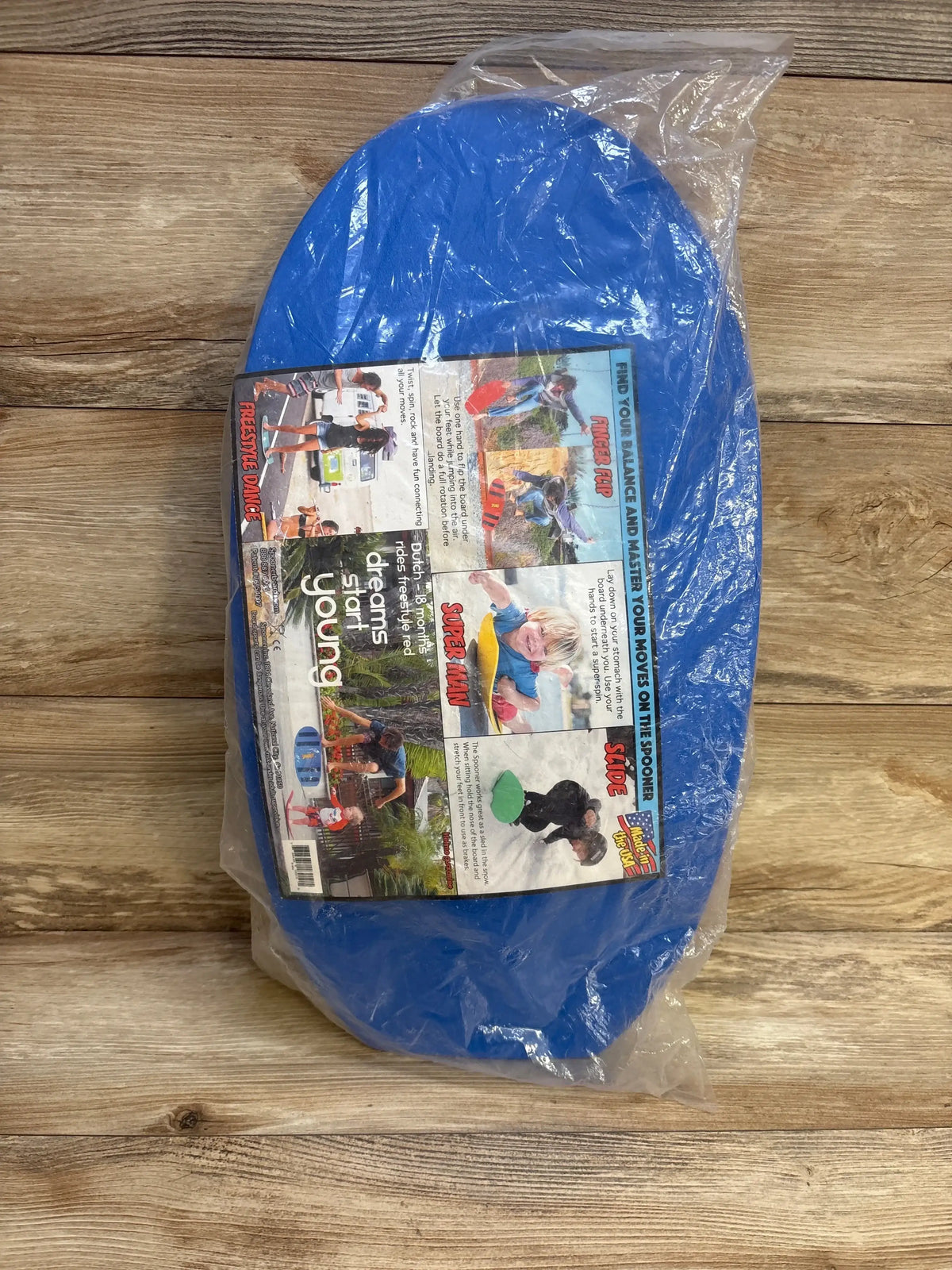 Angled top view of blue Spooner Board Pro in plastic packaging with product label showing 18m+ on wooden background