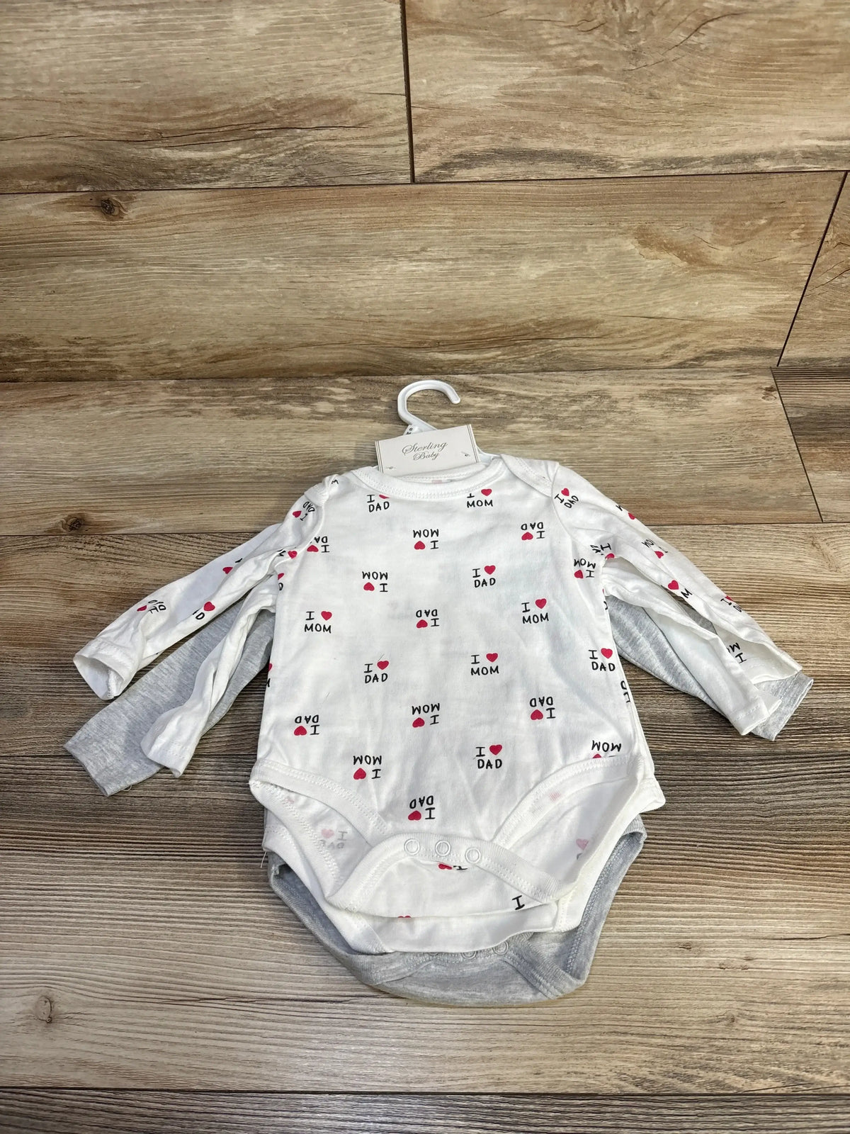 Front view of Sterling Baby 3-pack bodysuits showing long sleeves, lap shoulders, and snap crotch closures on a wood floor