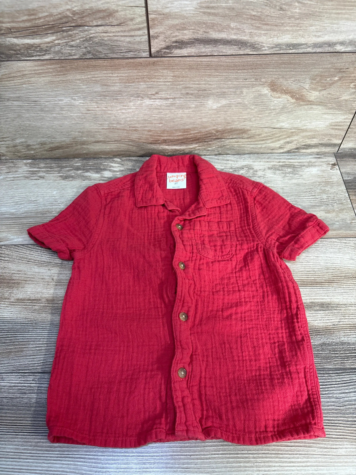 Front view of red Jumping Beans gauze button-up shirt, size 2T, with short sleeves and chest pocket on wood background