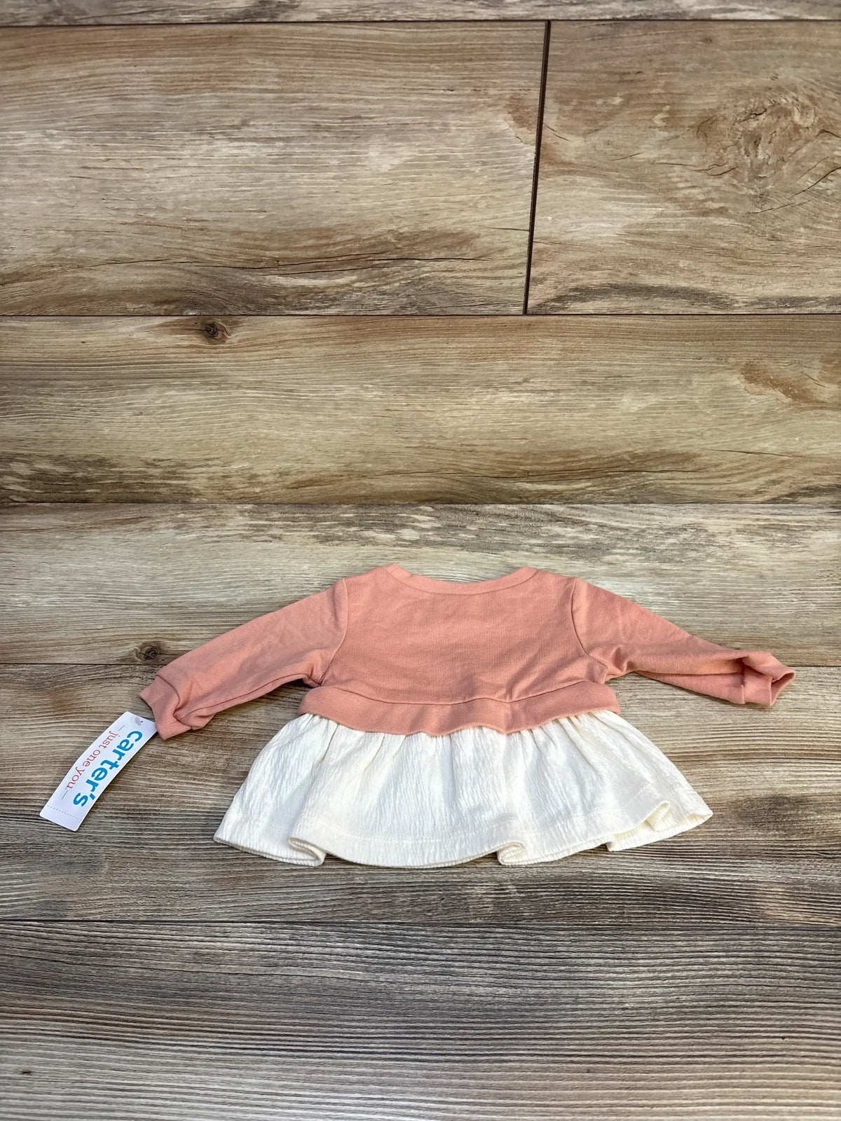 Flat lay of newborn peach peplum sweatshirt showcasing long sleeves and gathered white ruffle hem on wood floor background